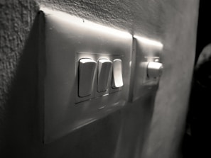 A close-up view of a wall-mounted electrical switch panel with three white switches and a dimly lit background. The surrounding wall has a slightly rough texture, and the scene is captured in grayscale, giving it a vintage feel.