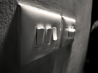 A close-up view of a wall-mounted electrical switch panel with three white switches and a dimly lit background. The surrounding wall has a slightly rough texture, and the scene is captured in grayscale, giving it a vintage feel.