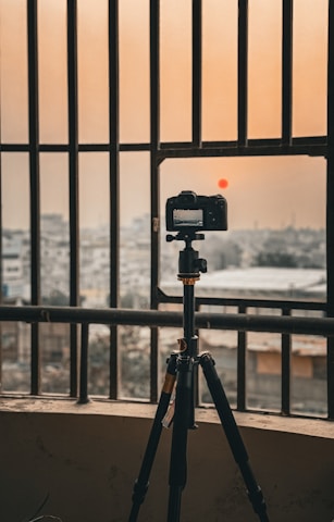 A camera mounted on a tripod stands in front of a balcony railing, overlooking a cityscape under a hazy orange sky. The sun is low in the sky, giving off a warm, diffused glow.
