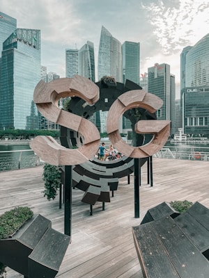 A wooden sculpture spelling out 'SG' on a waterfront platform, surrounded by skyscrapers and modern buildings. People can be seen near the sculpture, indicating a public space or tourist area. The sky is partly cloudy, providing a mix of natural and urban elements.