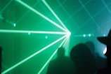 Green laser beams pierce through a foggy or smoky atmosphere, creating dynamic light patterns. Silhouettes of people are faintly visible in the background, contributing to a mysterious and immersive environment.