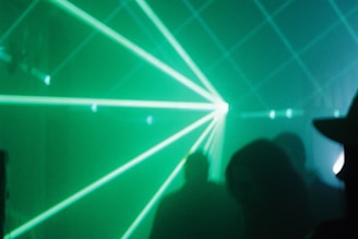 Green laser beams pierce through a foggy or smoky atmosphere, creating dynamic light patterns. Silhouettes of people are faintly visible in the background, contributing to a mysterious and immersive environment.