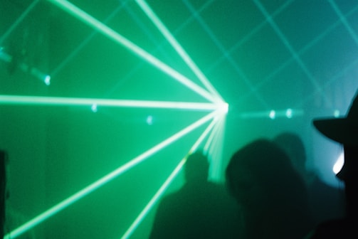 Green laser beams pierce through a foggy or smoky atmosphere, creating dynamic light patterns. Silhouettes of people are faintly visible in the background, contributing to a mysterious and immersive environment.