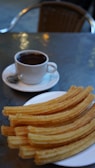 A cup of coffee sits next to a plate of churros on a metallic table. The coffee is in a white cup with a saucer, while the churros are golden and neatly arranged, suggesting a cozy and inviting atmosphere.
