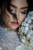 A close-up of a woman with her eyes closed, holding a bouquet of flowers near her face. Her makeup is soft and subtle, with a focus on her long eyelashes. Her hair is styled with loose strands falling naturally. The flowers are a mix of white and cream, complementing her sequined dress.