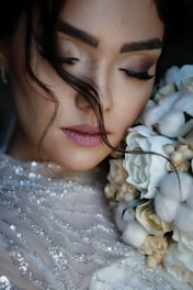 A close-up of a woman with her eyes closed, holding a bouquet of flowers near her face. Her makeup is soft and subtle, with a focus on her long eyelashes. Her hair is styled with loose strands falling naturally. The flowers are a mix of white and cream, complementing her sequined dress.