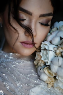 A close-up of a woman with her eyes closed, holding a bouquet of flowers near her face. Her makeup is soft and subtle, with a focus on her long eyelashes. Her hair is styled with loose strands falling naturally. The flowers are a mix of white and cream, complementing her sequined dress.