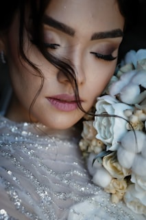 A close-up of a woman with her eyes closed, holding a bouquet of flowers near her face. Her makeup is soft and subtle, with a focus on her long eyelashes. Her hair is styled with loose strands falling naturally. The flowers are a mix of white and cream, complementing her sequined dress.