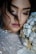 A close-up of a woman with her eyes closed, holding a bouquet of flowers near her face. Her makeup is soft and subtle, with a focus on her long eyelashes. Her hair is styled with loose strands falling naturally. The flowers are a mix of white and cream, complementing her sequined dress.