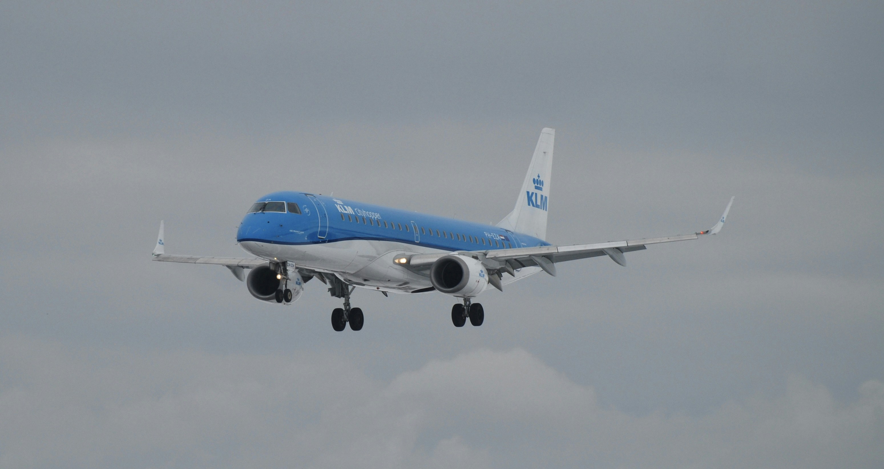 a blue and white airplane is flying in the sky, KLM Embraer landing in Gdańsk flying from Amsterdam in the snowy winter, taken with Nikon D80 and 70-300mm zoom lens also from Nikon