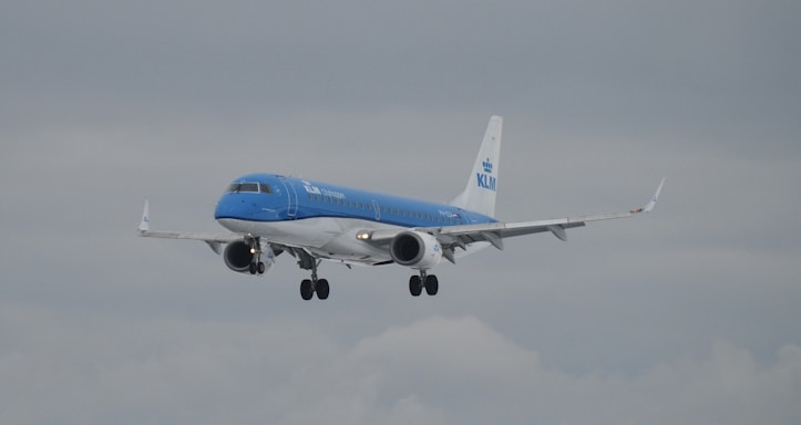 A commercial airplane in flight with a blue and white color scheme and KLM branding on the fuselage, against a backdrop of a cloudy sky.