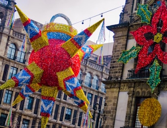 A vibrant, large pi&ntilde;ata with a spherical body and cone-shaped points is suspended in the air, decorated with colorful strips in red, yellow, blue, and green. It is set against a backdrop of historic architectural buildings with arched windows. A festive poinsettia and a circular decoration also hang alongside the pi&ntilde;ata.