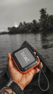 A person holding a small portable speaker with the brand name 'boAt'. The background features a serene water body and silhouettes of trees, suggesting a natural or outdoor setting.