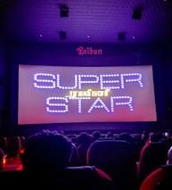 A dimly lit movie theater with an audience seated in front of a large screen displaying the words 'SUPER STAR' in bright, decorative text. The room is dark with subtle purple lighting, and there are audience heads silhouetted against the light from the screen.