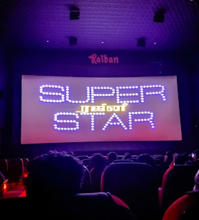 A dimly lit movie theater with an audience seated in front of a large screen displaying the words 'SUPER STAR' in bright, decorative text. The room is dark with subtle purple lighting, and there are audience heads silhouetted against the light from the screen.