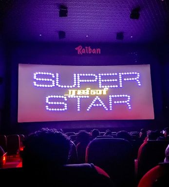 A dimly lit movie theater with an audience seated in front of a large screen displaying the words 'SUPER STAR' in bright, decorative text. The room is dark with subtle purple lighting, and there are audience heads silhouetted against the light from the screen.