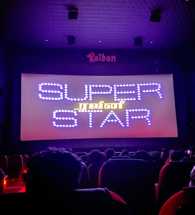 A dimly lit movie theater with an audience seated in front of a large screen displaying the words 'SUPER STAR' in bright, decorative text. The room is dark with subtle purple lighting, and there are audience heads silhouetted against the light from the screen.