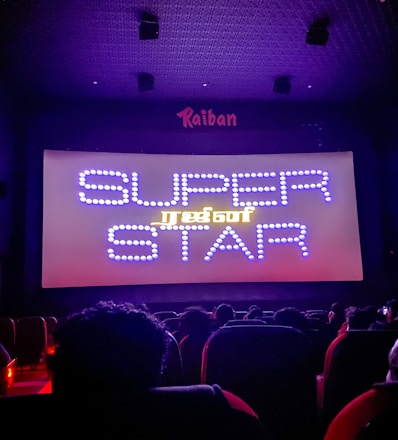 A dimly lit movie theater with an audience seated in front of a large screen displaying the words 'SUPER STAR' in bright, decorative text. The room is dark with subtle purple lighting, and there are audience heads silhouetted against the light from the screen.
