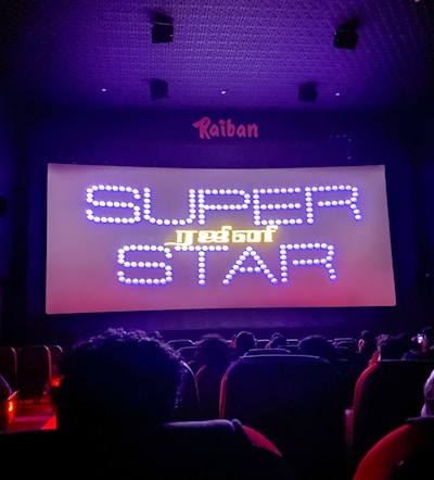 A dimly lit movie theater with an audience seated in front of a large screen displaying the words 'SUPER STAR' in bright, decorative text. The room is dark with subtle purple lighting, and there are audience heads silhouetted against the light from the screen.