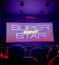A dimly lit movie theater with an audience seated in front of a large screen displaying the words 'SUPER STAR' in bright, decorative text. The room is dark with subtle purple lighting, and there are audience heads silhouetted against the light from the screen.