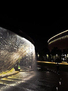 A nighttime scene featuring a cascading water feature illuminated by artificial lights, creating a shimmering effect. On the left, water flows over a curved structure, while a person sits on the wet steps nearby. In the background, trees and a cylindrical building with vertical lighting are visible.