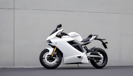 A sleek white motorcycle with modern aerodynamic design stands against a plain gray wall. Featuring sharp lines and a futuristic appearance, the motorcycle has black accents and a comfortable-looking seat. The tires are large with visible disc brakes, and the suspensions add to the sporty look.