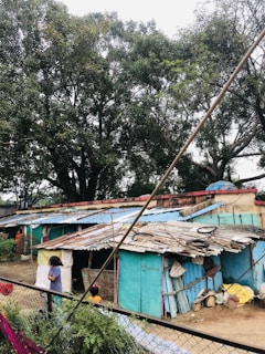A cluster of small, makeshift homes with corrugated metal roofs, some covered in blue and rusted patches. Tall trees with dense green foliage surround the area. Two children are visible, one standing near the corner of the structure, partially blocked from view. The ground is cluttered with various materials and bags, and a chain-link fence separates the scene from the foreground.