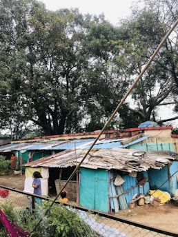 A cluster of small, makeshift homes with corrugated metal roofs, some covered in blue and rusted patches. Tall trees with dense green foliage surround the area. Two children are visible, one standing near the corner of the structure, partially blocked from view. The ground is cluttered with various materials and bags, and a chain-link fence separates the scene from the foreground.