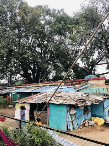 A cluster of small, makeshift homes with corrugated metal roofs, some covered in blue and rusted patches. Tall trees with dense green foliage surround the area. Two children are visible, one standing near the corner of the structure, partially blocked from view. The ground is cluttered with various materials and bags, and a chain-link fence separates the scene from the foreground.