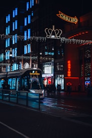 A bustling urban street scene at night featuring a tram heading to Centraal Station. The buildings are adorned with bright lights, including neon signs for a casino and Body Worlds exhibition. Christmas decorations hang above the street, adding a festive atmosphere. A group of people are seen walking along the sidewalk.
