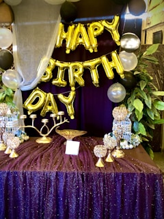 A decorative setup featuring gold balloon letters spelling out 'Happy Birthday' is prominently displayed. The background is adorned with a rich purple curtain, and the table in front is covered in a sparkling purple cloth. The table holds various golden decorative items, including a candelabra and globe-shaped ornaments. Surrounding the setup are lush green plants and helium balloons in black, white, and metallic colors.