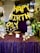 A decorative setup featuring gold balloon letters spelling out 'Happy Birthday' is prominently displayed. The background is adorned with a rich purple curtain, and the table in front is covered in a sparkling purple cloth. The table holds various golden decorative items, including a candelabra and globe-shaped ornaments. Surrounding the setup are lush green plants and helium balloons in black, white, and metallic colors.