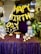 A decorative setup featuring gold balloon letters spelling out 'Happy Birthday' is prominently displayed. The background is adorned with a rich purple curtain, and the table in front is covered in a sparkling purple cloth. The table holds various golden decorative items, including a candelabra and globe-shaped ornaments. Surrounding the setup are lush green plants and helium balloons in black, white, and metallic colors.