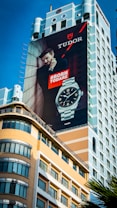 A large outdoor advertisement is displayed on the side of a tall building. The ad features a man leaning against a wall, looking confidently into the distance, with a prominent display of a wristwatch. The words 'Born to Dare' are boldly written beside him. The architectural style of the building includes a mix of modern elements and classical design.