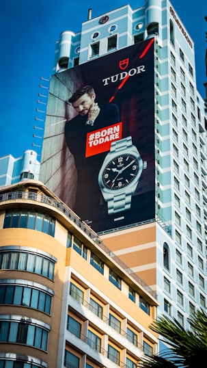 A large outdoor advertisement is displayed on the side of a tall building. The ad features a man leaning against a wall, looking confidently into the distance, with a prominent display of a wristwatch. The words 'Born to Dare' are boldly written beside him. The architectural style of the building includes a mix of modern elements and classical design.