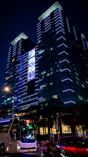 A modern, tall building is illuminated with bright blue lights. The structure features a large digital screen displaying content, adding a dynamic element to the facade. In the foreground, a bus and a motorcycle are visible, highlighting urban street activity at night. Trees and additional buildings can be seen in the lower portion of the scene.