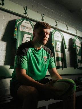 A rugby player sits on a bench in a locker room holding a rugby ball. He is wearing a green sports jersey, and behind him, jerseys numbered 6 and 8 are hanging on hooks against a brick wall. The room is dimly lit, creating a focused and contemplative atmosphere.