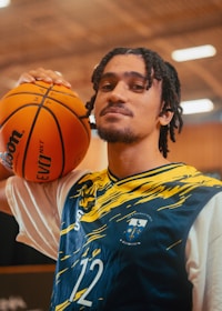 A person with shoulder-length braided hair is wearing a sports jersey with blue and yellow patterns and the number 12. They are holding a basketball on their shoulder. The background appears to be an indoor sports facility with blurred lighting.