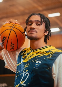 A person with shoulder-length braided hair is wearing a sports jersey with blue and yellow patterns and the number 12. They are holding a basketball on their shoulder. The background appears to be an indoor sports facility with blurred lighting.