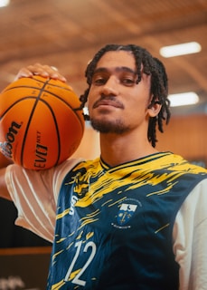A person with shoulder-length braided hair is wearing a sports jersey with blue and yellow patterns and the number 12. They are holding a basketball on their shoulder. The background appears to be an indoor sports facility with blurred lighting.