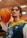 A person with shoulder-length braided hair is wearing a sports jersey with blue and yellow patterns and the number 12. They are holding a basketball on their shoulder. The background appears to be an indoor sports facility with blurred lighting.