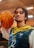 A person with shoulder-length braided hair is wearing a sports jersey with blue and yellow patterns and the number 12. They are holding a basketball on their shoulder. The background appears to be an indoor sports facility with blurred lighting.