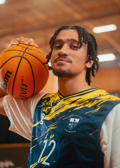 A person with shoulder-length braided hair is wearing a sports jersey with blue and yellow patterns and the number 12. They are holding a basketball on their shoulder. The background appears to be an indoor sports facility with blurred lighting.