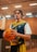 A basketball player stands in an indoor court holding a basketball. He is wearing a blue and yellow jersey with a unique design. The court has a wooden floor with lighting fixtures on the ceiling, and the background includes blurred stadium seating.