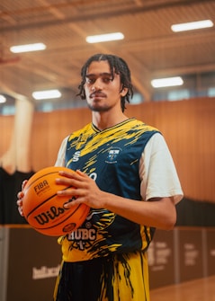 A basketball player stands in an indoor court holding a basketball. He is wearing a blue and yellow jersey with a unique design. The court has a wooden floor with lighting fixtures on the ceiling, and the background includes blurred stadium seating.