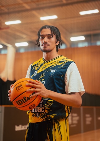 A basketball player stands in an indoor court holding a basketball. He is wearing a blue and yellow jersey with a unique design. The court has a wooden floor with lighting fixtures on the ceiling, and the background includes blurred stadium seating.