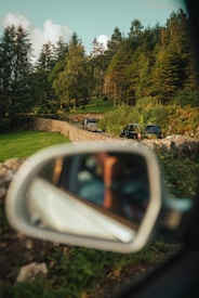 A serene forest scene with cars traveling along a narrow, winding road flanked by a stone wall. Tall, lush green trees and a clear blue sky create a peaceful, natural setting. The image includes a car's side mirror in the foreground, reflecting a blurred view of the driver.
