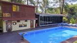 A serene poolside setting features a rectangular blue swimming pool bordered by wooden decking. Adjacent to the pool is a two-story wooden structure with large windows and a smaller section with artistic murals and a door. The area is surrounded by lush green trees and comfortable seating creates a relaxing environment.