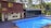 A serene poolside setting features a rectangular blue swimming pool bordered by wooden decking. Adjacent to the pool is a two-story wooden structure with large windows and a smaller section with artistic murals and a door. The area is surrounded by lush green trees and comfortable seating creates a relaxing environment.