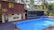 A serene poolside setting features a rectangular blue swimming pool bordered by wooden decking. Adjacent to the pool is a two-story wooden structure with large windows and a smaller section with artistic murals and a door. The area is surrounded by lush green trees and comfortable seating creates a relaxing environment.